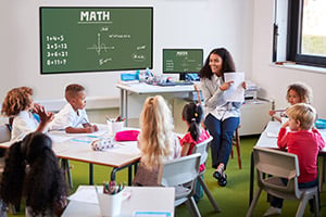 Smiling female infant school teacher sitting on a chair facing school kids in a classroom holding up and explaining a worksheet to them