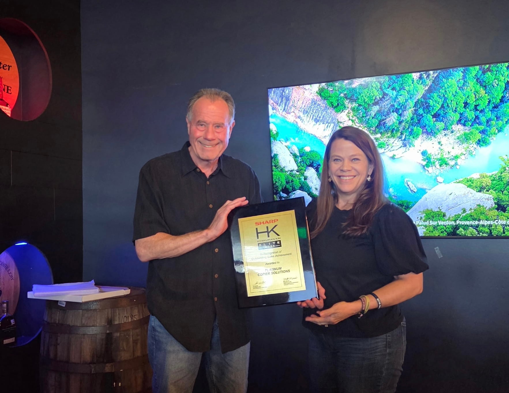 Two Adults Stand Side by Side Indoors Smiling Holding a Framed Award Plaque Together During a Presentation
