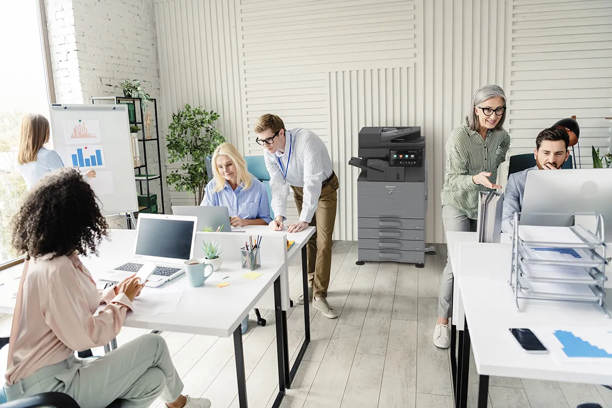 How to Estimate Copier Lease Cost for Your Business Six coworkers collaborate at bright white desks in a modern office; charts on a board and a copier in the background.