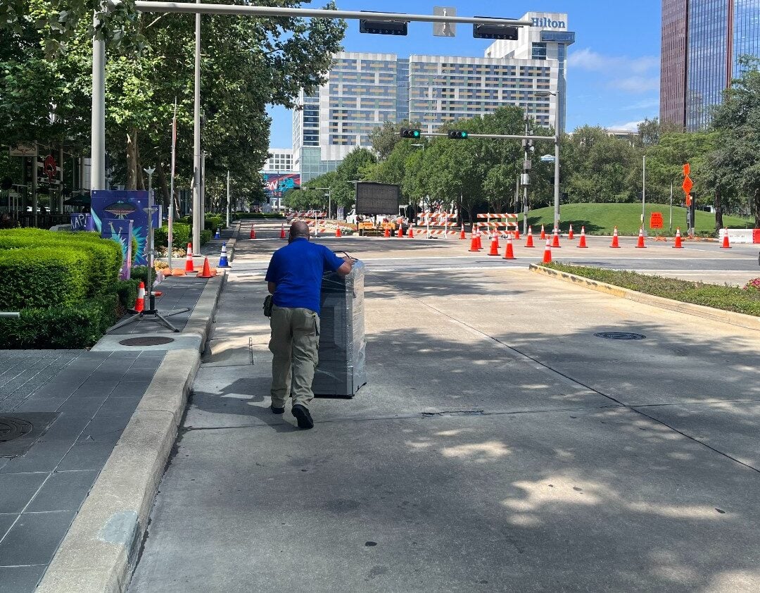 5f01f2eb 4465 45e9 897e fc546ba3fad1 | platinum copier solutions Construction Worker in a Blue Shirt Pushes a Large Gray Metal Cabinet Along a Closed City Street Orange Traffic Cones Lining the Road with a Hilton Hotel in the Background