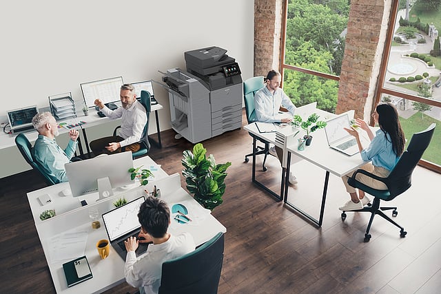 Managed Print Services Benefits for Business Efficiency Above view of business employees at desks in a bright office setting with a Sharp printer in the background.