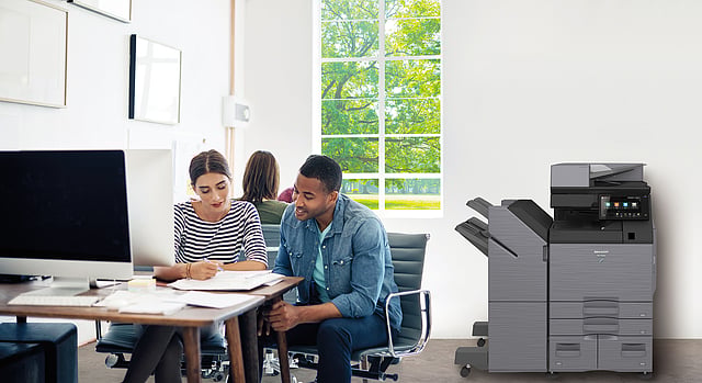 How Does the Copier Leasing Process Work for Businesses? Two coworkers sit at a desk reviewing papers, with a large gray multifunction printer in a bright office corner.