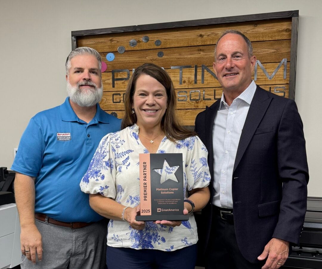 Three people pose in an office; woman in the center holds a Platinum Copier Solutions award plaque, flanked by two men in business attire.