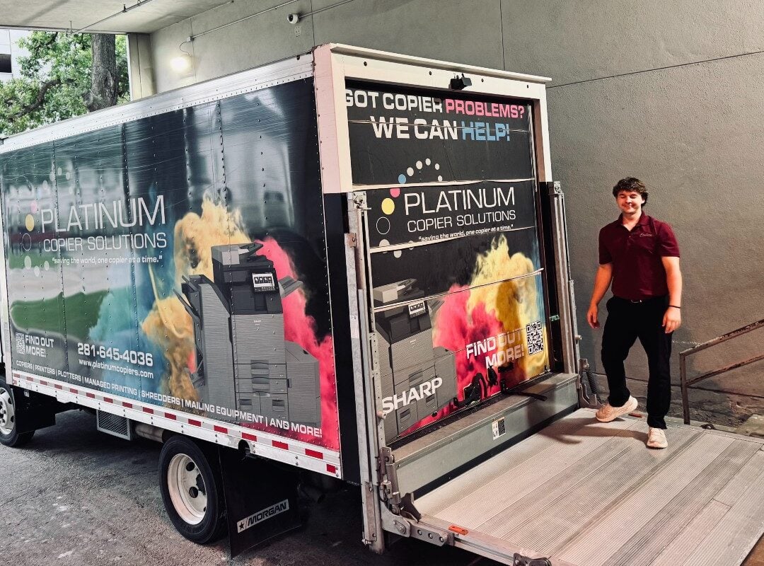 Advertising moving truck for Platinum Copier Solutions with colorful printer imagery; man in maroon polo stands at open rear ramp.