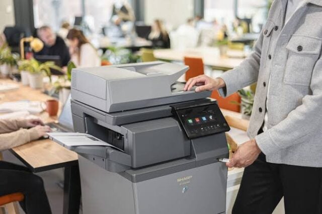 | platinum copier solutions Person Using a Large Multifunction Printercopier with a Touchscreen Panel in a Busy Open plan Office Space