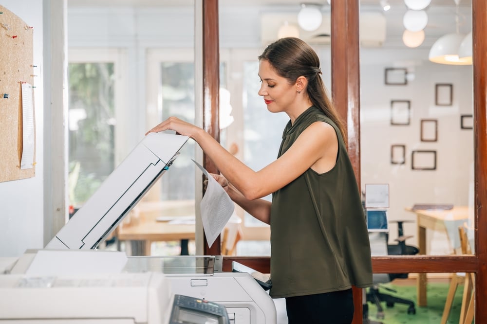 Print Cost Reduction Strategies with Managed Print Services Woman in a sleeveless olive-green top feeds paper into a photocopier in a bright office environment with a glass partition behind her.