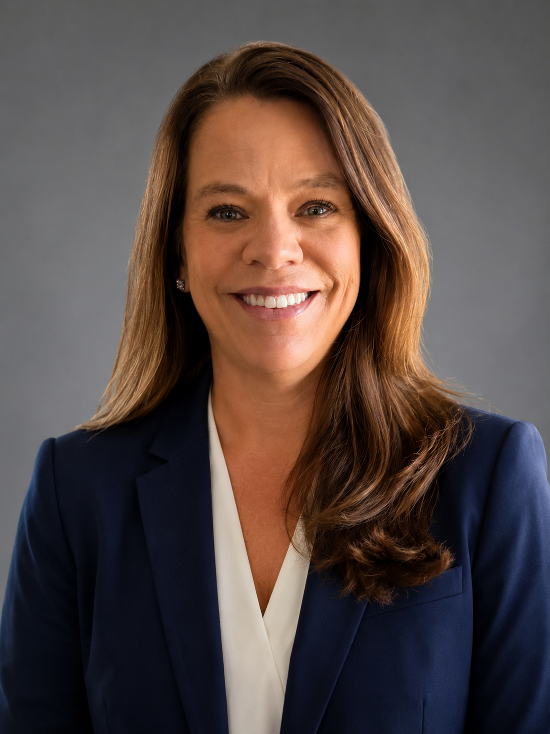 Professional headshot: smiling woman in a navy blazer and white blouse against a gray backdrop.