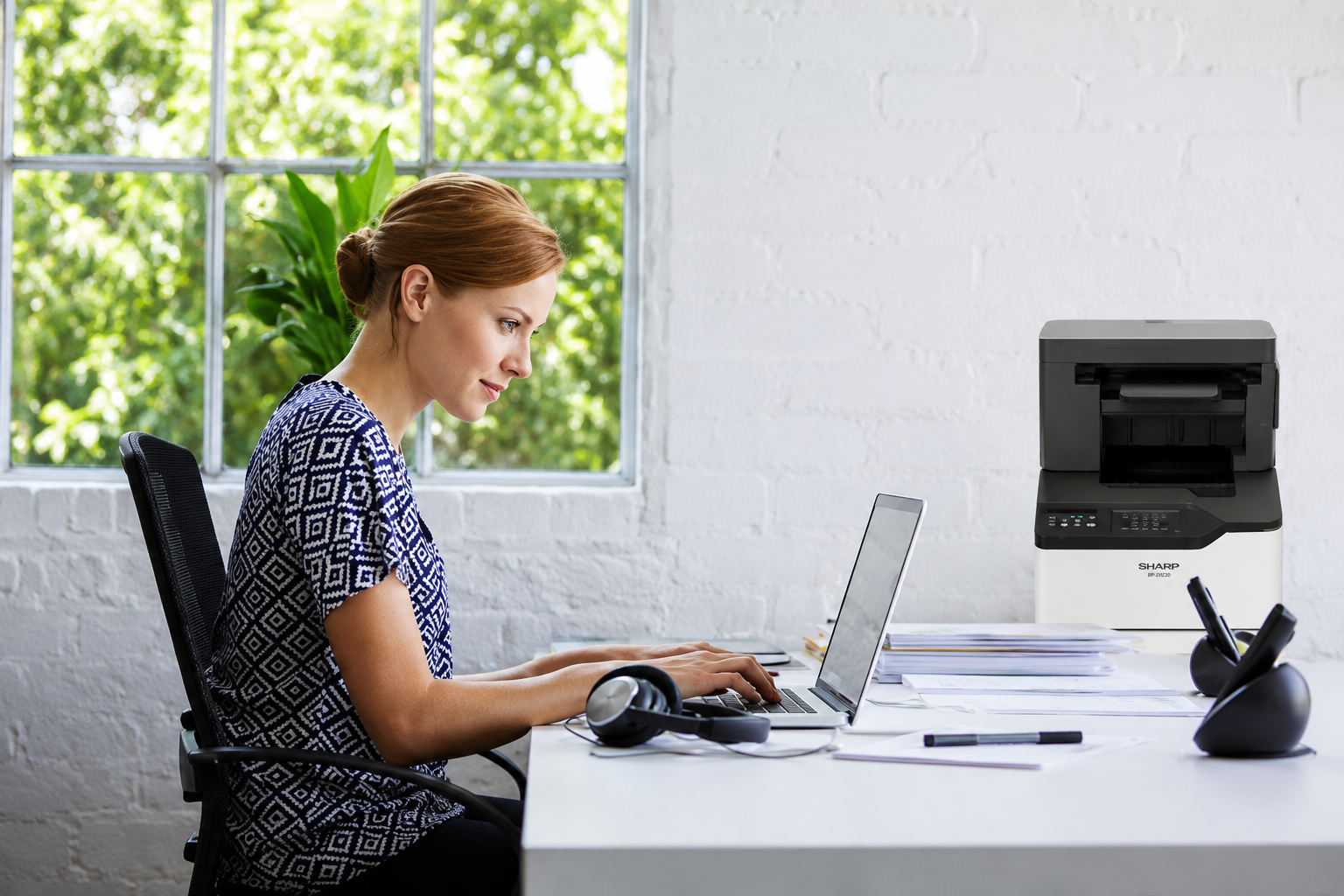 Woman working on a laptop at a bright white desk, with a printer and papers nearby by the window.