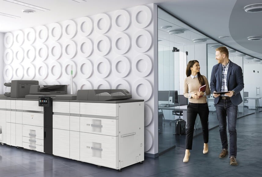 Managed Print Service Process: How It Works for Your Business Modern office lobby with a large multifunction printer at a white cabinet and two professionals walking and talking by a glass-walled meeting room, holding a tablet and a notebook.