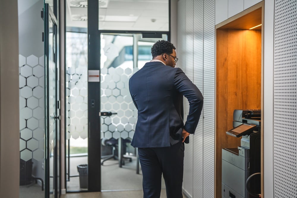 Printer Fleet Management: What Businesses Should Know African american businessman standing in an modern office corridor, waiting by a multifunction printer to collect printed or copied documents, representing productivity and work tasks.