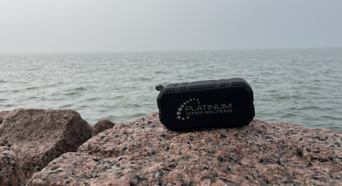Rugged Black Portable Speaker with Platinum Computer Solutions Logo Sits on Pink Granite Rocks by the Sea